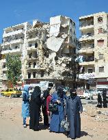 Women wait for bus near damaged building in Syria
